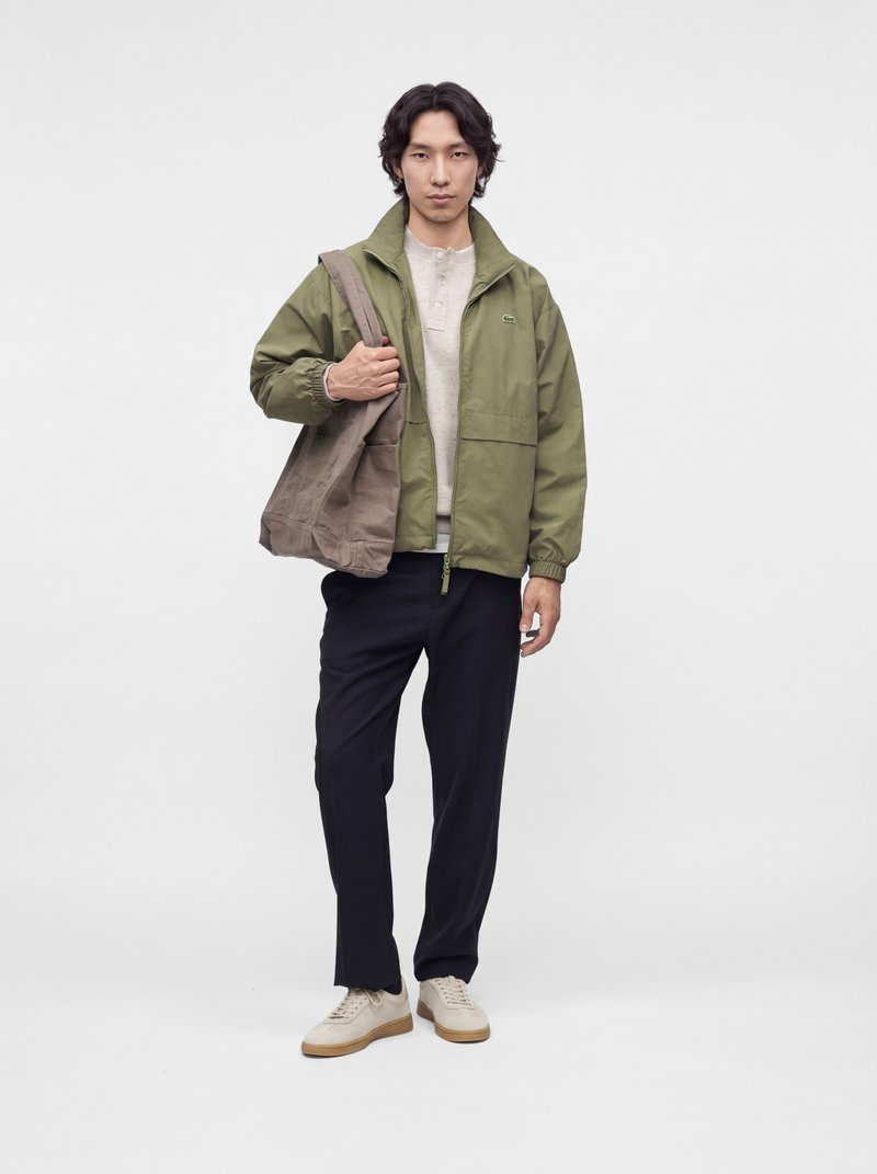 Jeune homme portant une veste verte, une chemise beige clair, un pantalon foncé, des baskets blanches, tenant un grand sac cabas marron sur l'épaule.