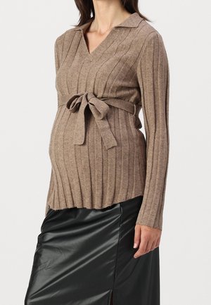 Gravid kvinde iført en brun ribstrikket sweater med et bundet bælte og et sort satinskørt med frontslids.