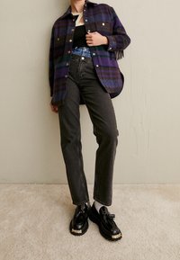 Chaqueta de cuadros en púrpura, negro y verde con detalles de flecos, combinada con pantalones rectos negros y mocasines de plataforma negros.