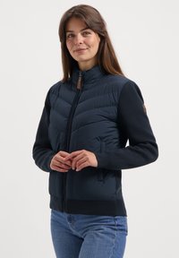 Jonge vrouw in een marineblauwe gewatteerde jas met gebreide mouwen en een blauwe spijkerbroek, staand tegen een effen lichte achtergrond, met een lichte glimlach.