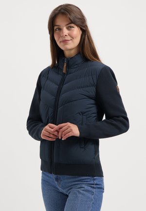 Jonge vrouw in een marineblauwe gewatteerde jas met gebreide mouwen en een blauwe spijkerbroek, staand tegen een effen lichte achtergrond, met een lichte glimlach.