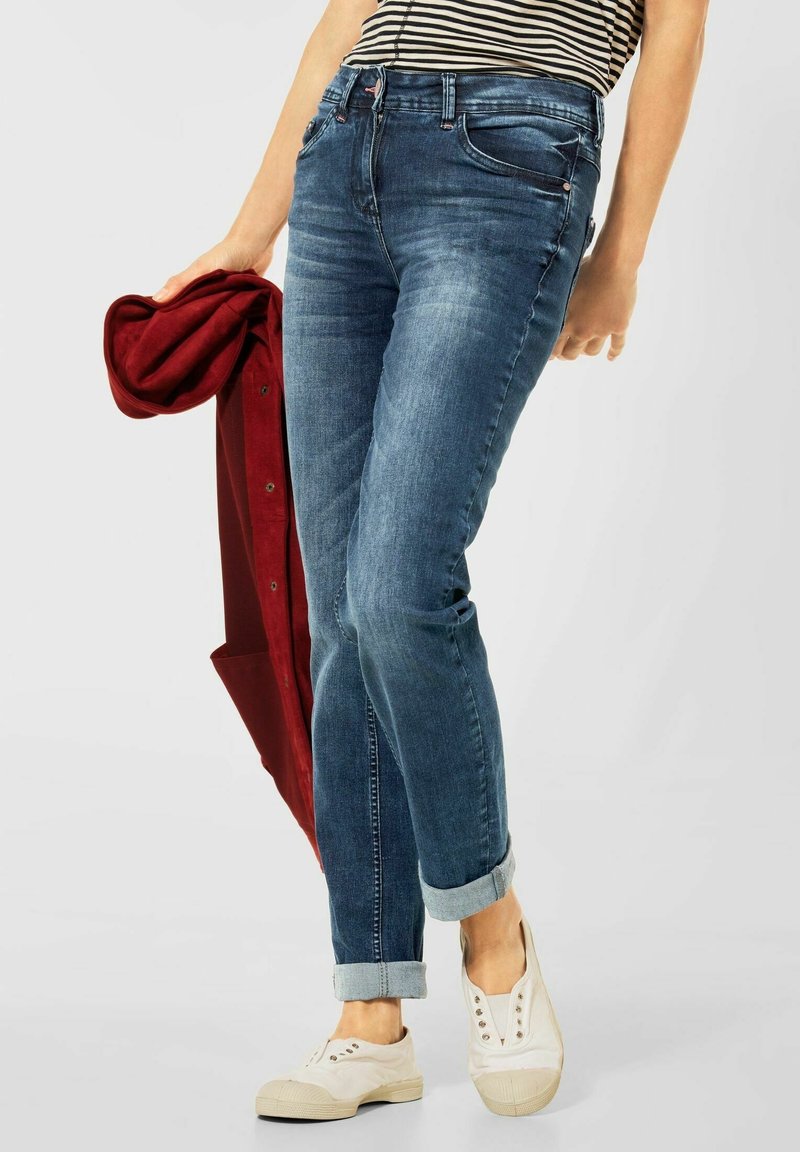 Personne portant un jean bleu retroussé et des chaussures blanches sans lacets, tenant une veste rouge pliée, chemise rayée visible au niveau de la taille.
