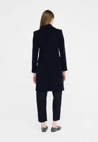 Marineblauwe wollen jas met een getailleerde silhouet, lange mouwen en een opvallend gestructureerde revers, gedragen over bijpassende cropped broek en platte schoenen.