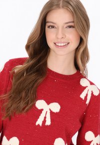 Maglione rosso lavorato a maglia decorato con motivi a fiocco bianchi e sottili luccichii. Presenta un collo tondo e maniche lunghe.