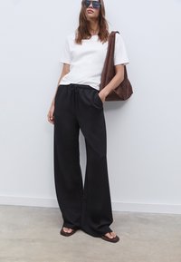 Zwarte wijde broek met elastische tailleband, gecombineerd met een witte shirt met korte mouwen, een bruine suède tote bag en donkere zonnebril.