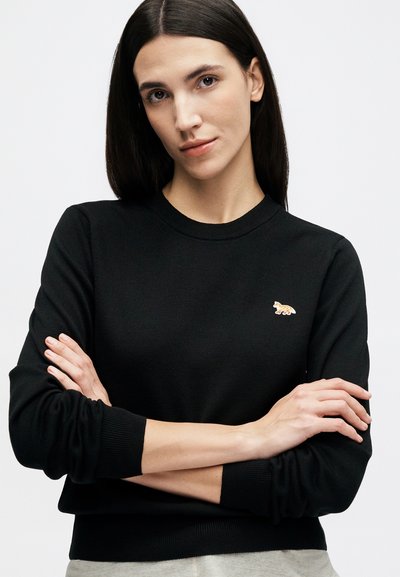 Γυναίκα με ίσια σκούρα μαλλιά που φοράει μαύρο πουκάμισο με μακριά μανίκια και ένα μικρό έμβλημα αλεπούς στο στήθος, με σταυρωμένα τα χέρια.