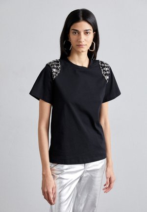 T-shirt nera con maniche corte, caratterizzata da decorazioni e dettagli lavorati sulle spalle. Abbinata a pantaloni lucidi argentati.