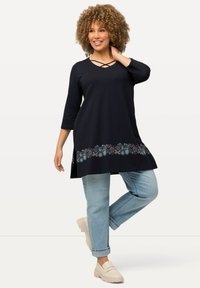 Marineblaues Tunika mit crisscross-Ausschnitt und blumischer Stickerei am Saum. Getragen mit hellblauen, hochgekrempelten Jeans und beigen Plateauschuhen.