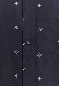 Camicia button-up navy con motivi geometrici bianchi e azzurri chiari. Presenta un pulsante nero e una texture di tessuto liscia.
