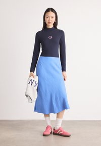 Marineblå langermet topp med logo, parret med en lys blå, utsvingt midiskjørt. Modellen holder en grå hettejakke, iført rosa sneakers og hvite sokker.