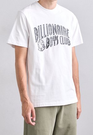 Mężczyzna noszący białą koszulkę z szarym napisem „Billionaire Boys Club” i grafiką hełmu astronauty, w połączeniu z jasnzielonymi spodniami.