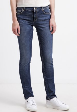 Personne portant un jean slim bleu foncé et des baskets blanches, debout devant un fond clair uni.