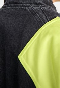 Närbild av axel på svart denimjacka med ett stygnat neon gult tygmärke på ärmen.