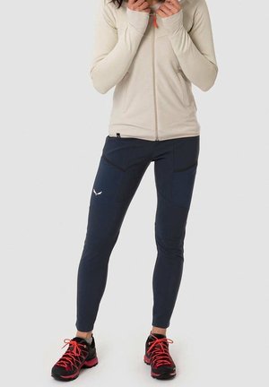 Kvinde iført en beige jakke med lynlås, navyblå tætsiddende bukser med lommer og sorte trail-løbesko med røde snørebånd, stående på en hvid baggrund.