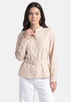 Kvinde med mørkt hår iført beige skjorte med bælte og hvide bukser, poserer med den ene hånd nær kraven mod en ensfarvet baggrund.