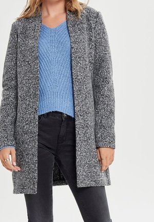 Donna che indossa un maglione azzurro a maglia con scollo a V, un cappotto grigio strutturato aperto davanti e jeans neri a vita alta, su uno sfondo semplice.