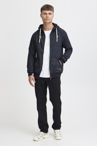 Gray zip-up hoodie with drawstring, contrasting dark accents, side pockets, and a logo detail. Paired with black pants and sneakers.