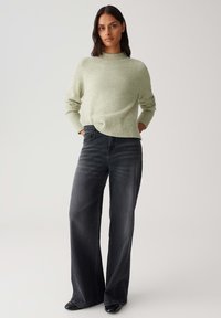 Woman stands with hands in pockets wearing light green sweater, black wide-leg jeans, and black shoes against plain background.