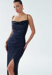Navy blue sequined dress with a cowl neckline, slim fit, and side slit. Features spaghetti straps and textured fabric for added visual interest.