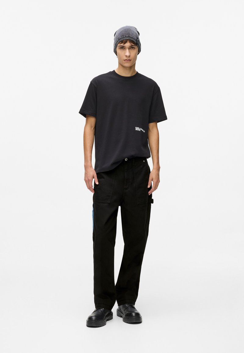 Pantaloni cargo negri cu accente albastre pe laterale, asortați cu un tricou negru lejer. Modelul poartă o șapcă gri și pantofi negri.