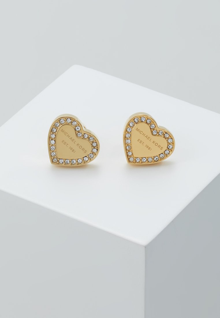 Orecchini a forma di cuore in oro con accenti di cristallo e incisione "MICHAEL KORS EST. 1981" su una superficie liscia e lucida.