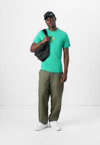 Grøn bomulds t-shirt med et gult grafik, parret med olivenfarvede cargo bukser og en sort crossbody taske. Hvide sneakers fuldender outfittet.