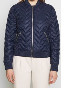 Tmavě modrá prošívaná bomber bunda s vzorem chevron, žebrovaným límcem a manžetami, předním zipovým zapínáním a dvěma zipovými postranními kapsami.
