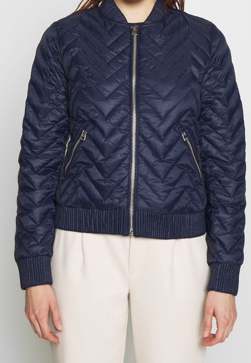 Tmavě modrá prošívaná bomber bunda s vzorem chevron, žebrovaným límcem a manžetami, předním zipovým zapínáním a dvěma zipovými postranními kapsami.