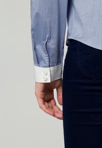 Personne portant une chemise bleue avec des boutons blancs aux poignets et un pantalon bleu foncé, main détendue sur le côté contre un fond uni.