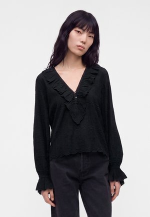 Vrouw met lang zwart haar draagt een zwarte blouse met lange mouwen en een ruches V-hals en zwarte broek, staand tegen een effen lichte achtergrond.