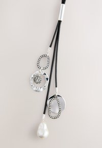 Schwarze Kordelhalskette mit mehreren Metallanhängern: silberne und schwarze Ringe, eine funkelnde Kugel und ein weißer Perlenanhänger. Strukturierte und polierte Oberflächen.