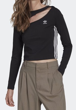 Femme portant un haut court noir Adidas à manches longues avec des bandes blanches sur les manches et un pantalon plissé marron taille haute.
