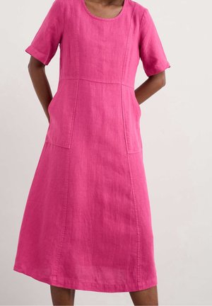 Kvinde iført en knælange, lyserød kjole med korte ærmer og lommer foran, stående foran en ensfarvet lys baggrund.