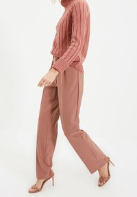 Rippene, hellrosa Rollkragensweatshirt kombiniert mit passenden Hosen, beide aus einem weichen Stoff. Braune, mit Riemchen ausgestattete High-Heels runden das Outfit ab.