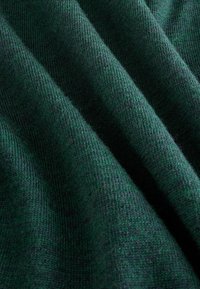 Tumši zaļš adīts audums ar redzamu austuma tekstūru un mīkstiem krokām, kas veido ēnas un gaismas atspīdējumus.