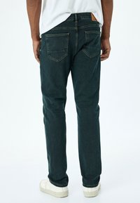 Jeans de mezclilla verde oscuro con perneras ligeramente ajustadas, que cuentan con dos bolsillos traseros y costuras en contraste. Se llevan con zapatos blancos.
