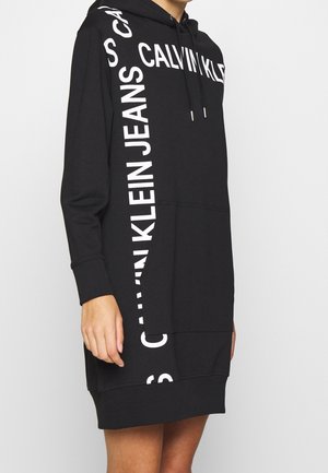 Osoba nosząca czarną długą sukienkę z kapturem typu sweatshirt z białym napisem Calvin Klein Jeans na piersi i boku.