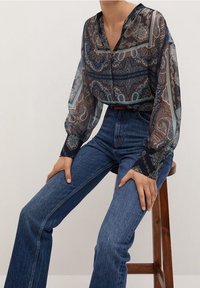 Blouse bleu marine transparent à motif cachemire, manches longues avec poignets foncés, portée avec un jean flare bleu taille haute.