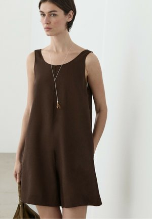 Frau trägt ein ärmelloses braunes Kleid und eine lange silberne Halskette mit Anhänger, hält eine braune Tasche in einer neutralen Innenumgebung.