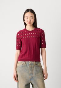 Kastanienbraunes gestricktes Oberteil mit kurzen Ärmeln, das horizontale Rippen und runde Ausschnitte aufweist. Kombiniert mit hellblauen Jeans.