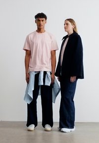 Άνδρας με ροζ μπλουζάκι και σκούρα παντελόνια, με ριγέ πουκάμισο δεμένο γύρω από τη μέση, γυναίκα με μαύρο μπουφάν και σκούρα τζιν με φαρδιά πόδια, στέκονται μπροστά σε απλό φόντο.