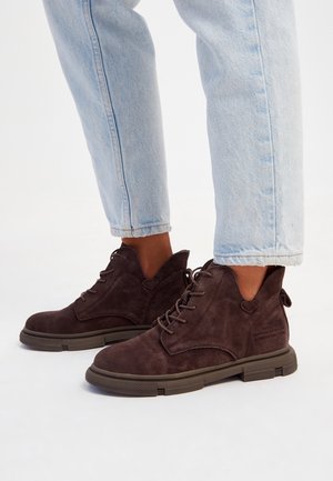 Bottes cheville en daim brun foncé avec lacets portées avec un jean bleu clair retroussé sur un fond blanc.