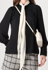 Czarna bluzka z koronkowymi detalami na ramionach, długimi rękawami i zapinana na guziki z przodu, zestawiona z białym szalikiem i kraciastą spódnicą tweedową.