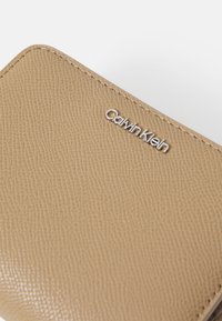 Billetera de cuero texturizado beige de Calvin Klein con acabado suave, que presenta un acento de logotipo plateado y esquinas redondeadas.
