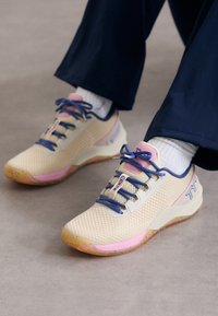Beżowe buty sportowe z siateczkową cholewką, różowymi i niebieskimi akcentami oraz teksturowaną gumową podeszwą. Posiadają wyraźne sznurowanie i detal z logo.