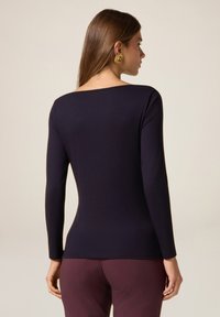 Top navy a maniche lunghe con scollatura quadrata, forma aderente, texture liscia, abbinato a pantaloni bordeaux. Orecchini dorati accentuano il look.
