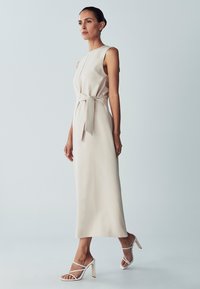 Sleeveless, A-line dress in soft beige fabric with a tied waist detail. Paired with white strappy heels featuring a gold accent.