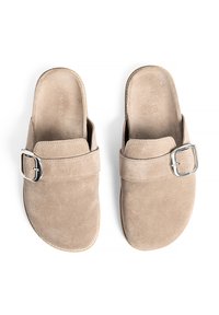 Beige krāsas zamšādas slip-on kuriem ir plata siksna ar sudraba sprādzi. Līdzena zole un noapaļoti pirksti, ar gludu tekstūru.