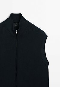 Svart ärmlös stickad väst med hög krage, med en dragkedja framtill och ribbad textur. Tillverkad av 100 % bomull. Varumärkesetikett synlig.