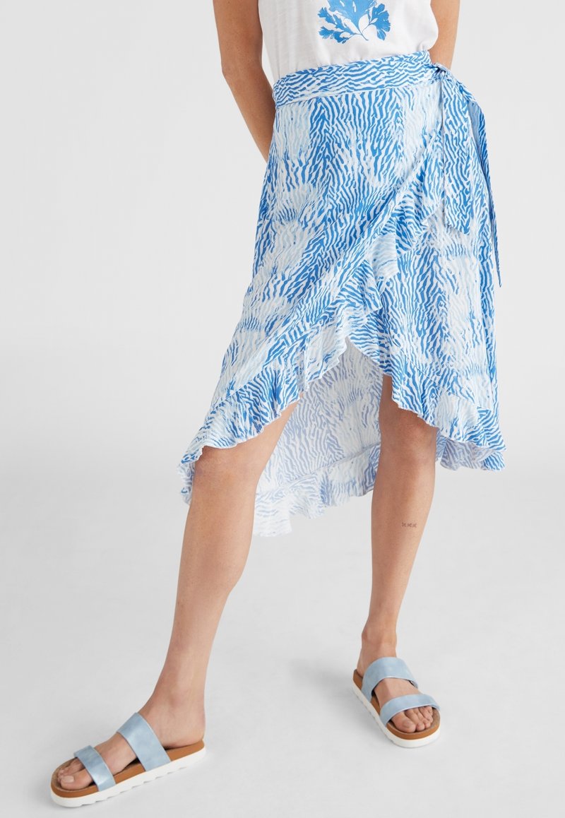 Femme portant une jupe portefeuille bleu et blanc avec des bords à volants, associée à des sandales légères bleu clair et une chemise blanche avec un imprimé de fleurs bleues.